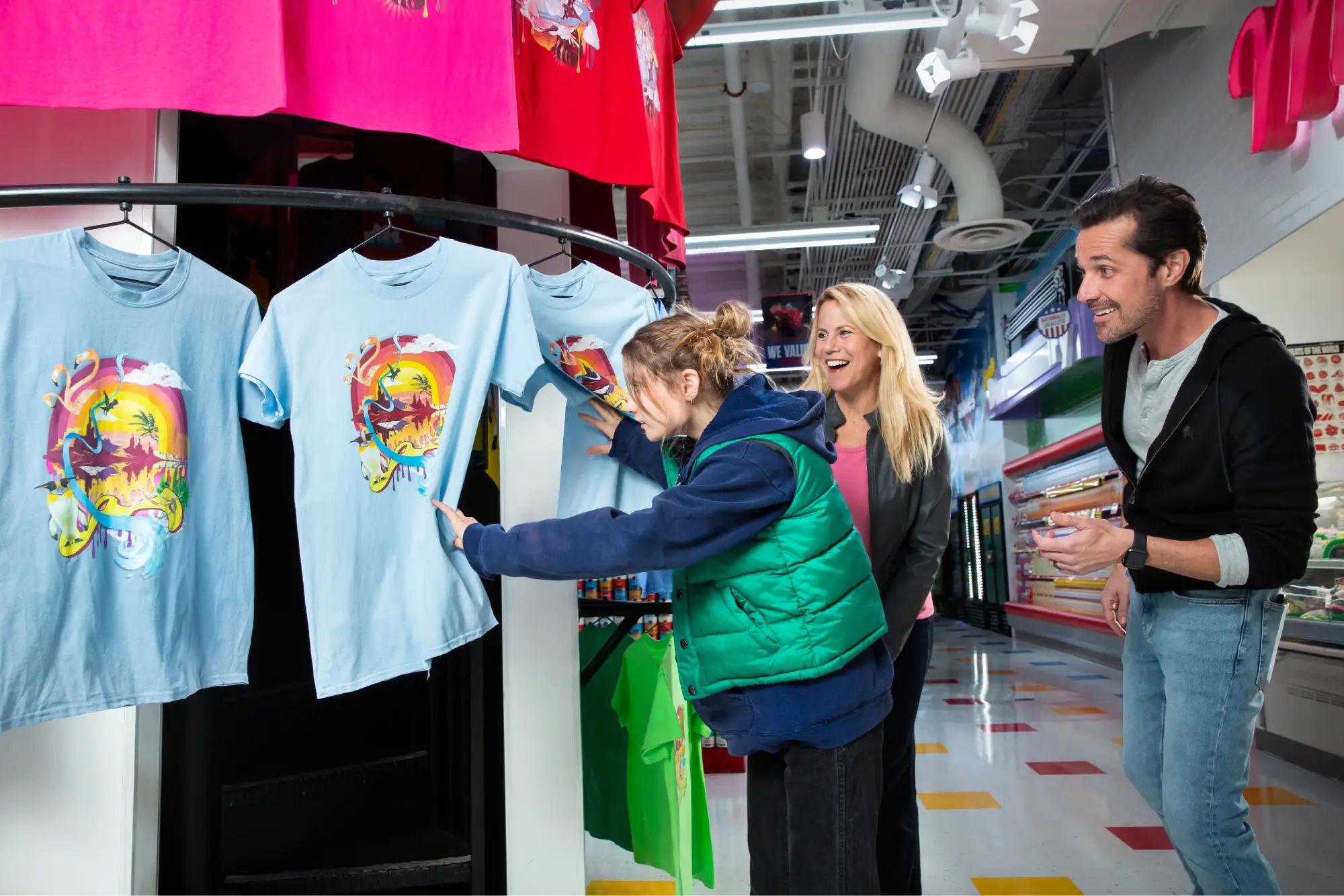 A young girl examines a blue graphic T-shirt while two adults watch her in a store aisle.
