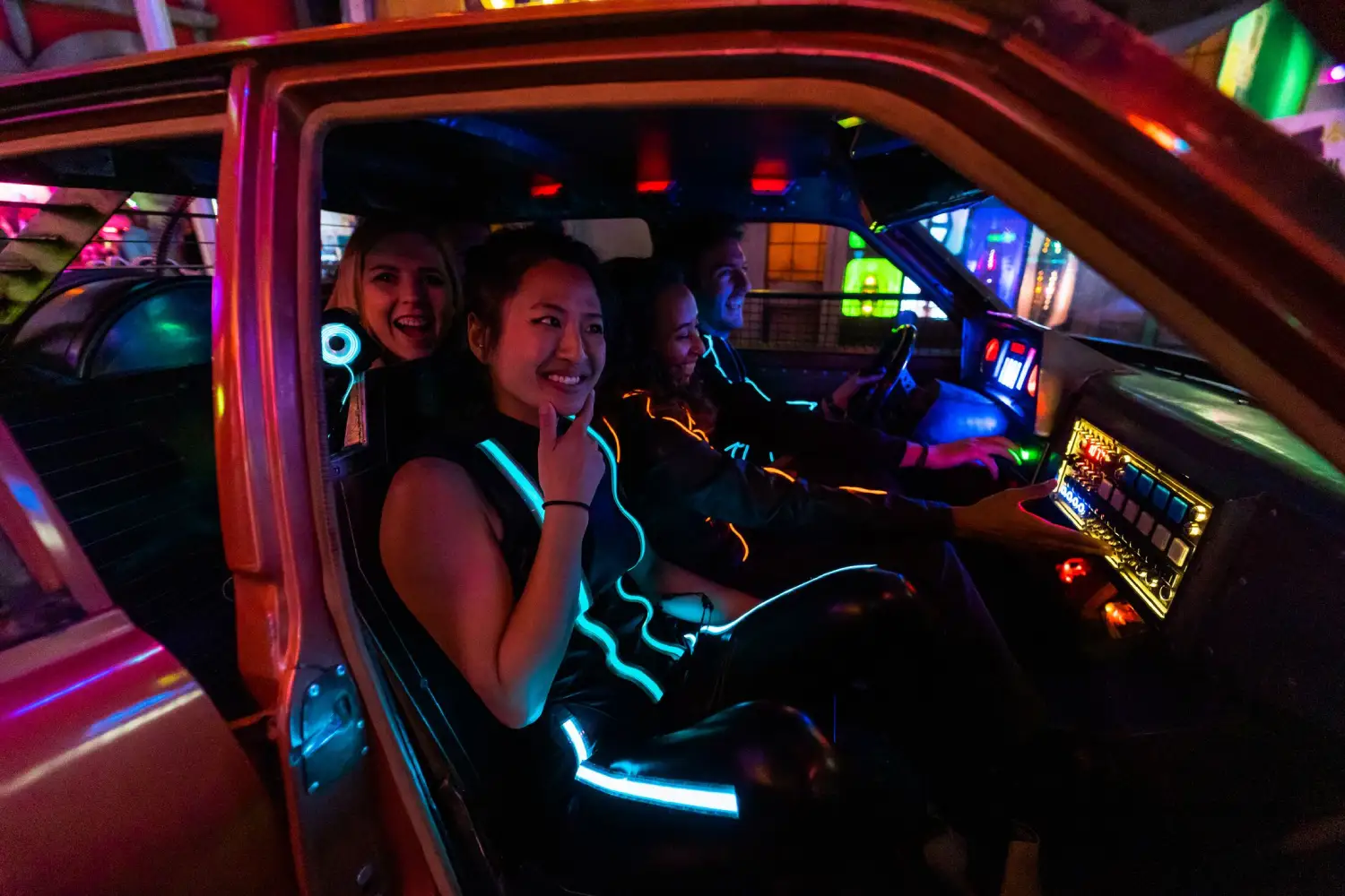 Guests sitting in neon car at Meow Wolf Denver’s convergence station immersive art experience, a storytelling experience of art inside this cosmic amusement center in denver.