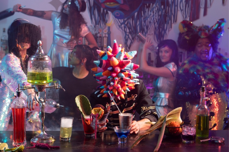 A group of people and a pointy head character have drinks at a bar.