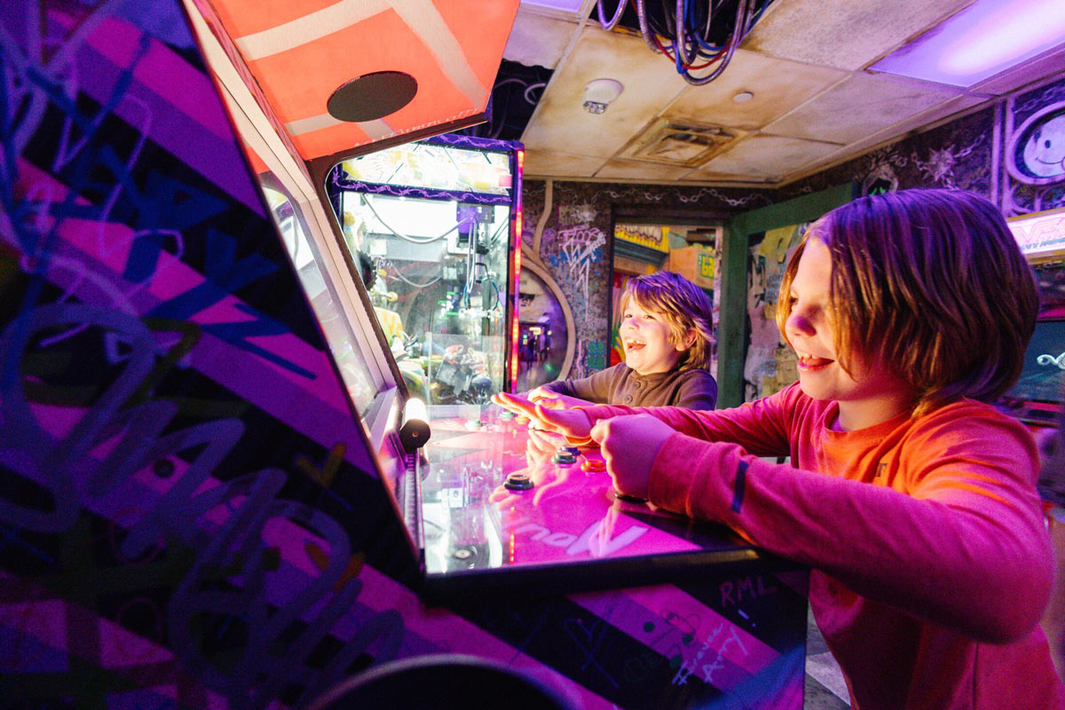 Two kids laugh and play side-by-side on a glowing pink arcade machine in a vibrant, graffiti-covered room at Meow Wolf Grapevine, surrounded by neon lights and immersive retro-futuristic decor.