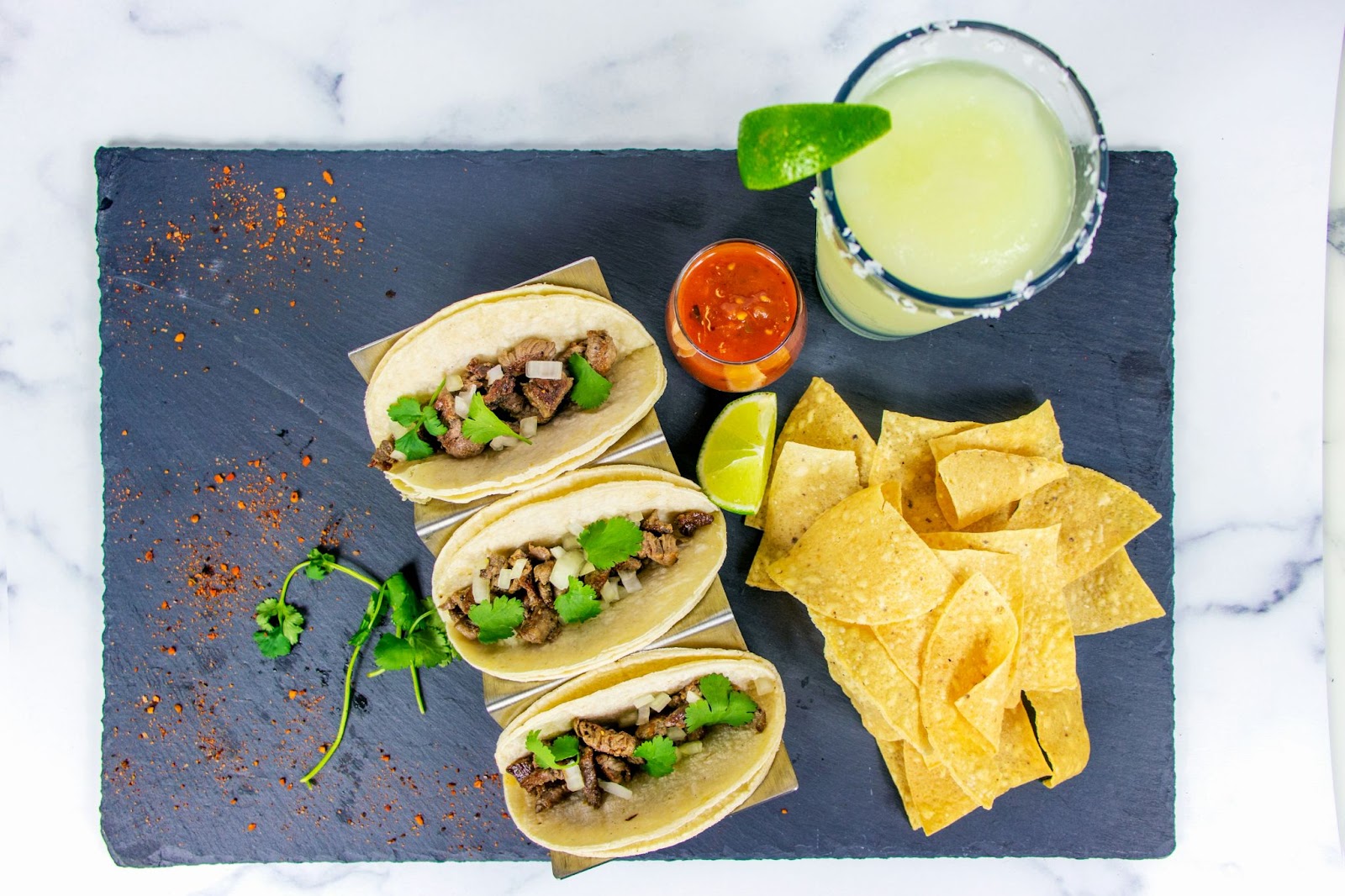 three tacos on a slate with chips, salsa and a margarita