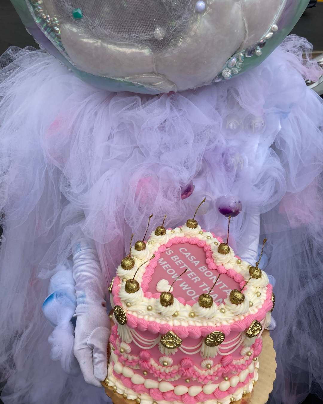 A fluffy Meow Wolf character holds a pink, white, and gold cake that says “Casa Bonita is Better Than Meow Wolf.”