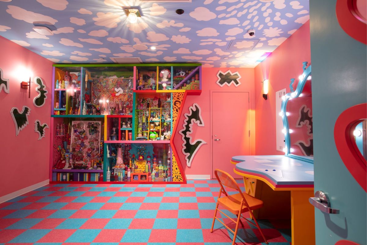 A pink room with a dresser and lights to the right and a bookshelf in the back with knickknacks. 