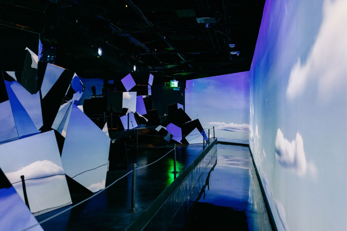 A room with projected walls and mirrors showing a cloudy sky.