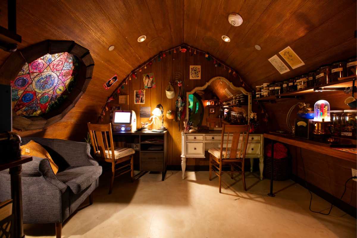 The interior of Rose & Marin’s dwelling features a stained glass window, a desk with a working computer, and various personal items from the residents’ lives, including books, unusual glowing fixtures, and herbs in jars.