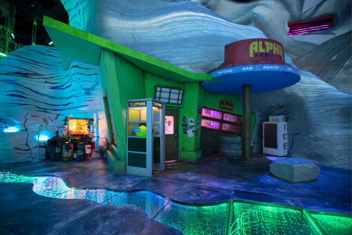 An empty gas station sits across a strange river of shifting light. A telephone booth and an ice machine are nearby.