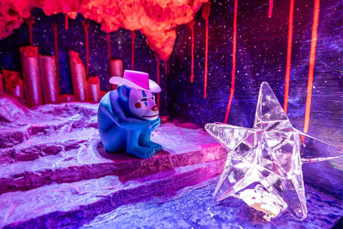 A blue creature with a clown mask and cowboy hat looks at a star shape in a rocky space.
