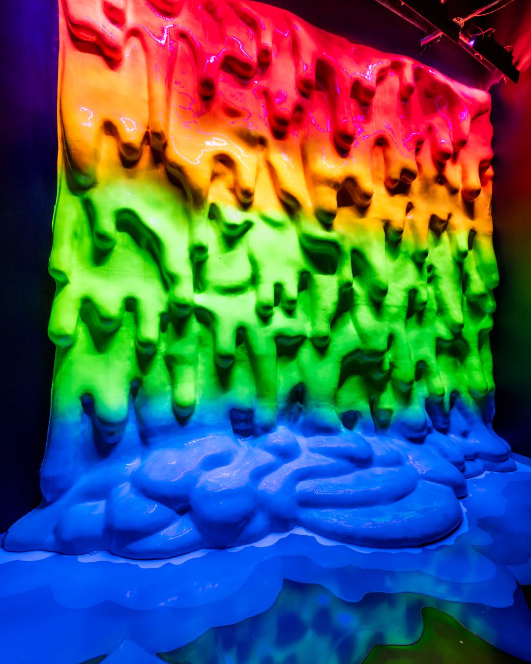 A rainbow colored dripping wall of paint with a pool of blue at the bottom.
