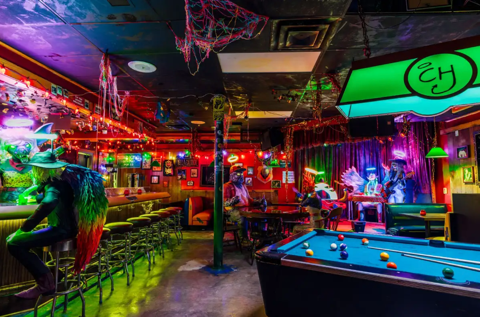 A bar to the left with a being with rainbow wings sitting at it, a pool table to the right, and a band of creatures on the stage.