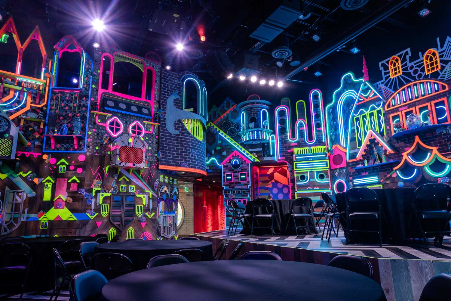 Tables and chairs are set up on the floor and stage of a room with neon lights on brick, house-looking fronts.