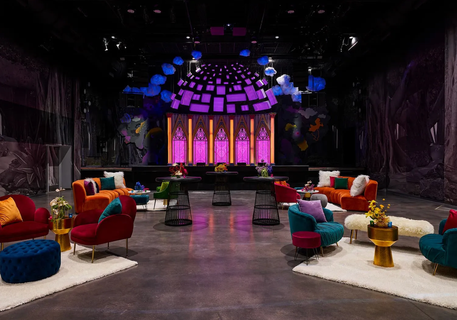 A large room with colorful tables, chairs, and rugs sits in-front of a purple stage with stained glass looking panes.