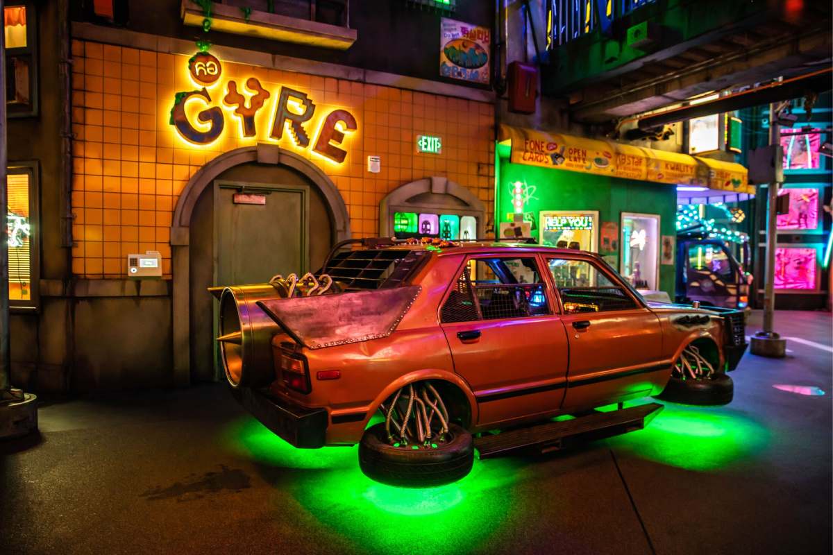 A floating red car with green lights underneath sits in front of a building with a “GYRE” sign above a doorway.