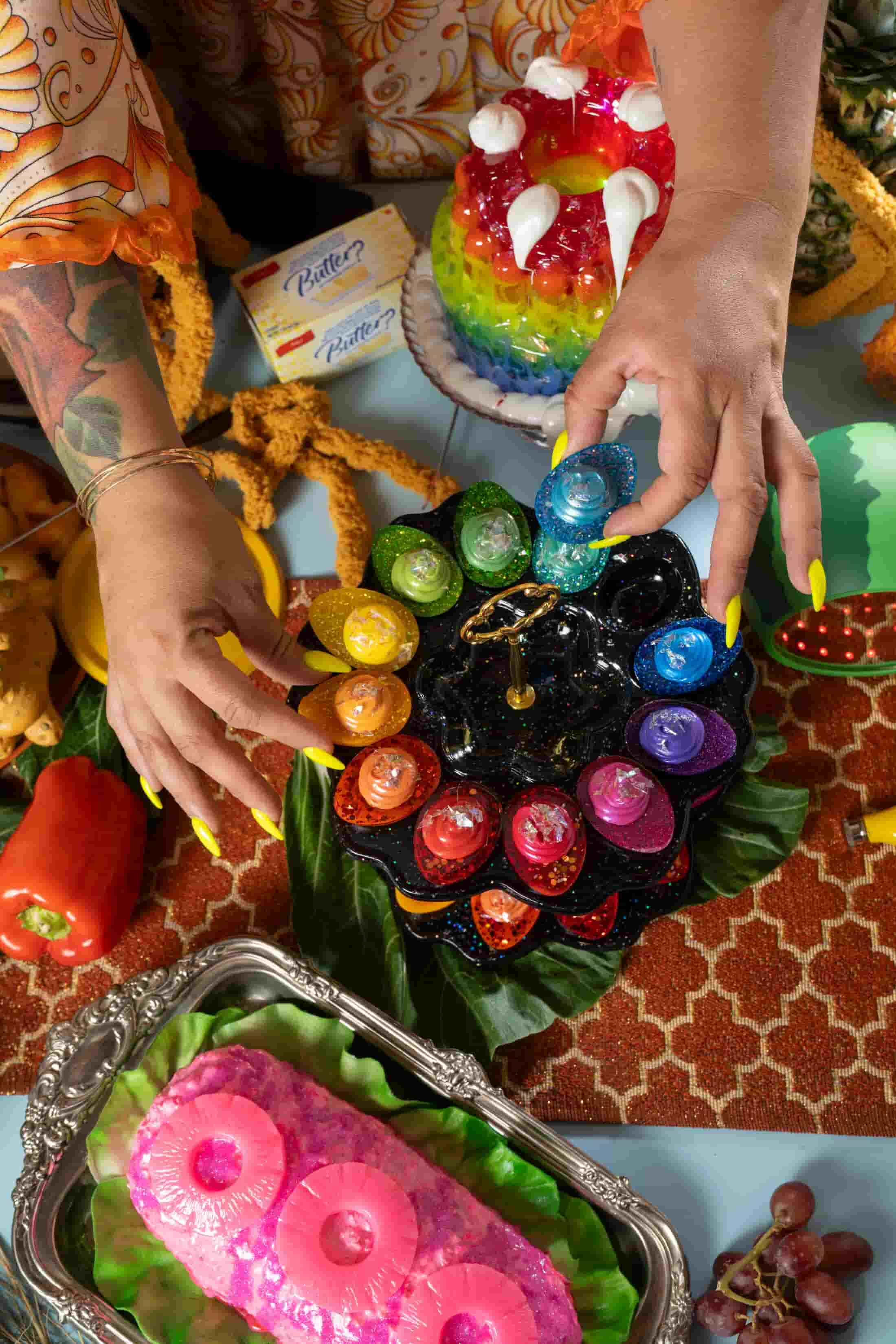 Hands with long yellow nails hold a rainbow deviled egg over an elaborate dinner table, with a neon pink meatloaf and a rainbow gelatin.