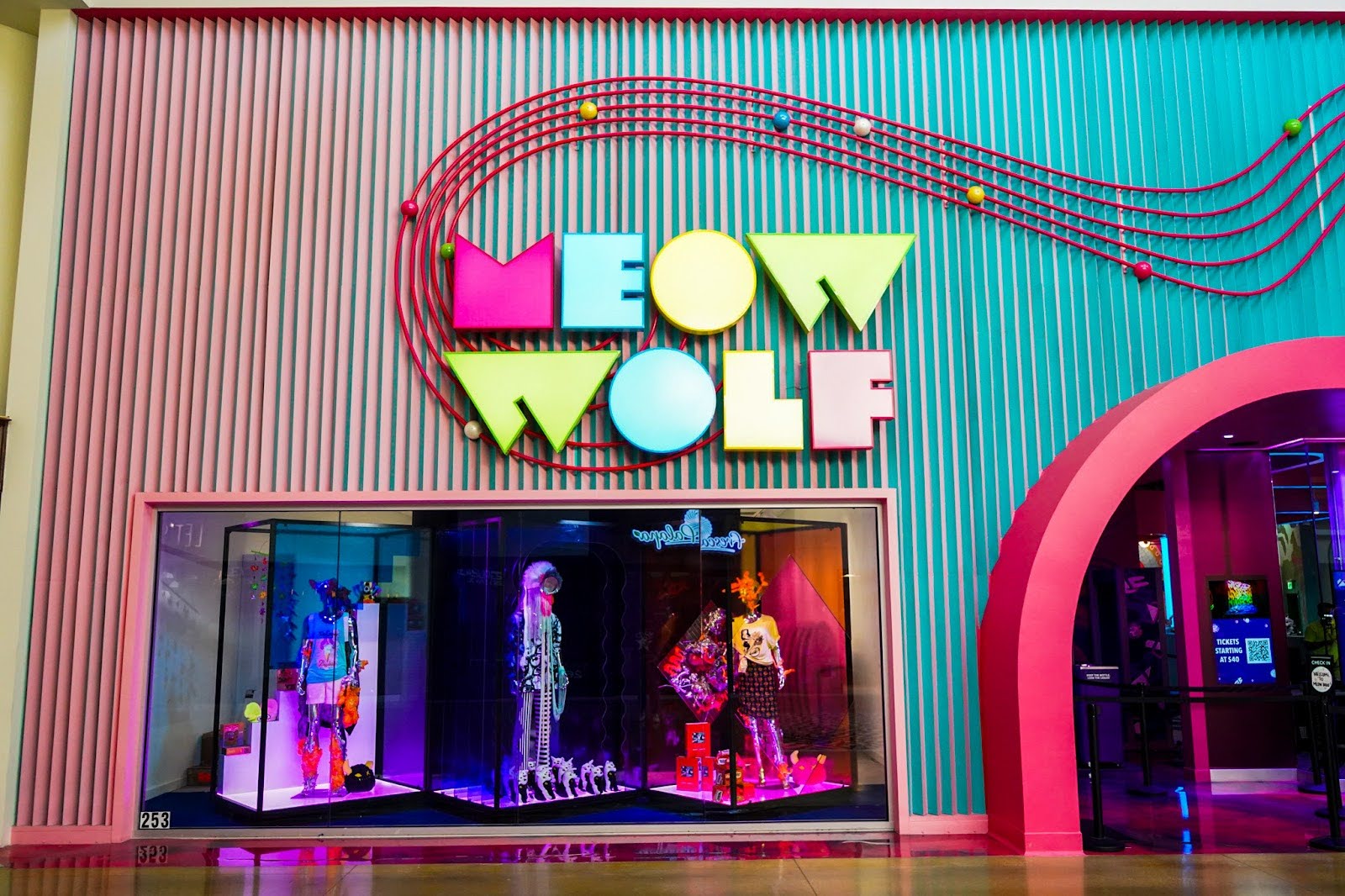 The interior mall storefront of Meow Wolf with panels switching color from pink to blue, and a display of mannequins in Meow Wolf merch
