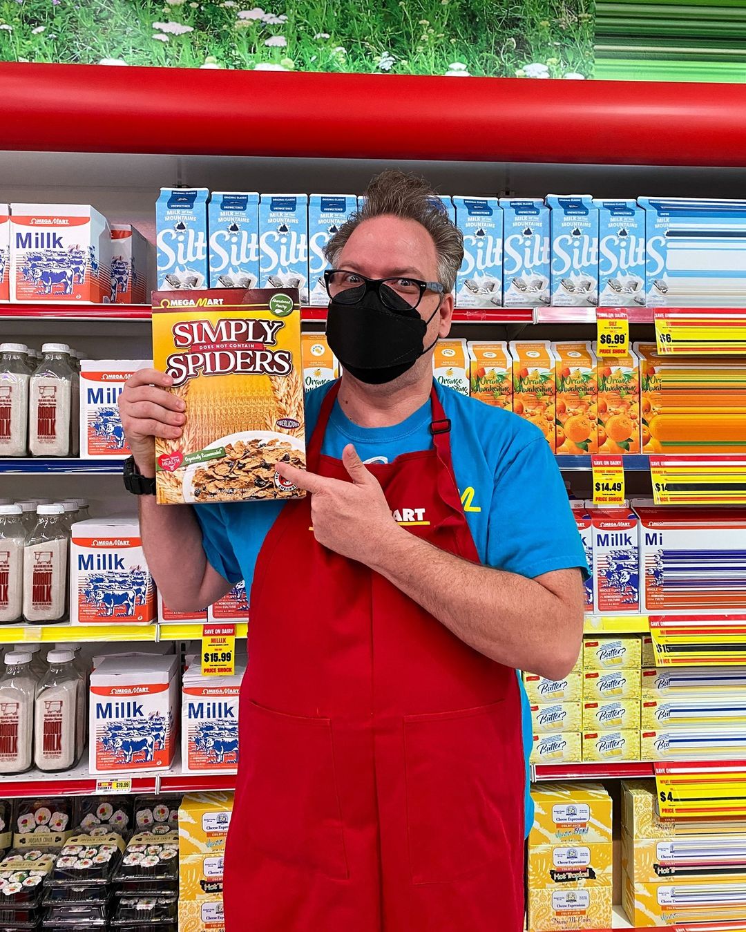 Omega Mart employee with a black mask and glasses, holding a box of Simply Spiders cereal and pointing to it