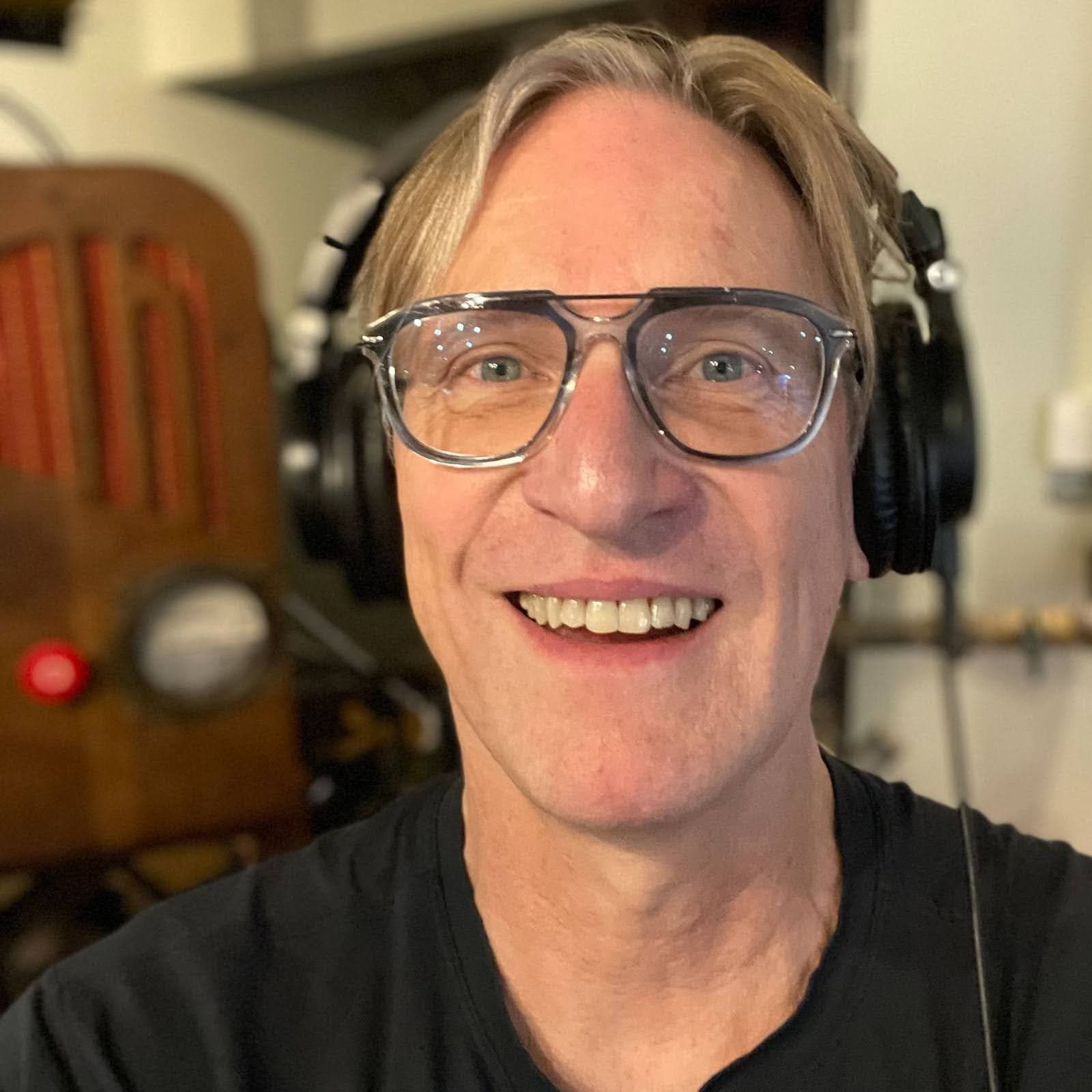 Headshot of Les (a white cis male) smiling and wearing glasses and headphones.