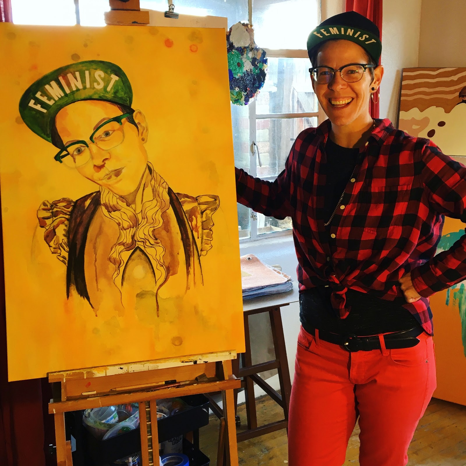 Jaelah Kuehmichel wearing a hat with "Feminist" on the underside of it standing next to a WIP painting