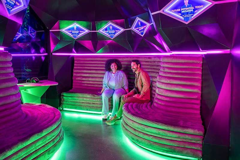 Two people sit on curved, cushioned benches inside a futuristic, neon-lit room with purple and green lighting, geometric walls, and digital screens glowing overhead.