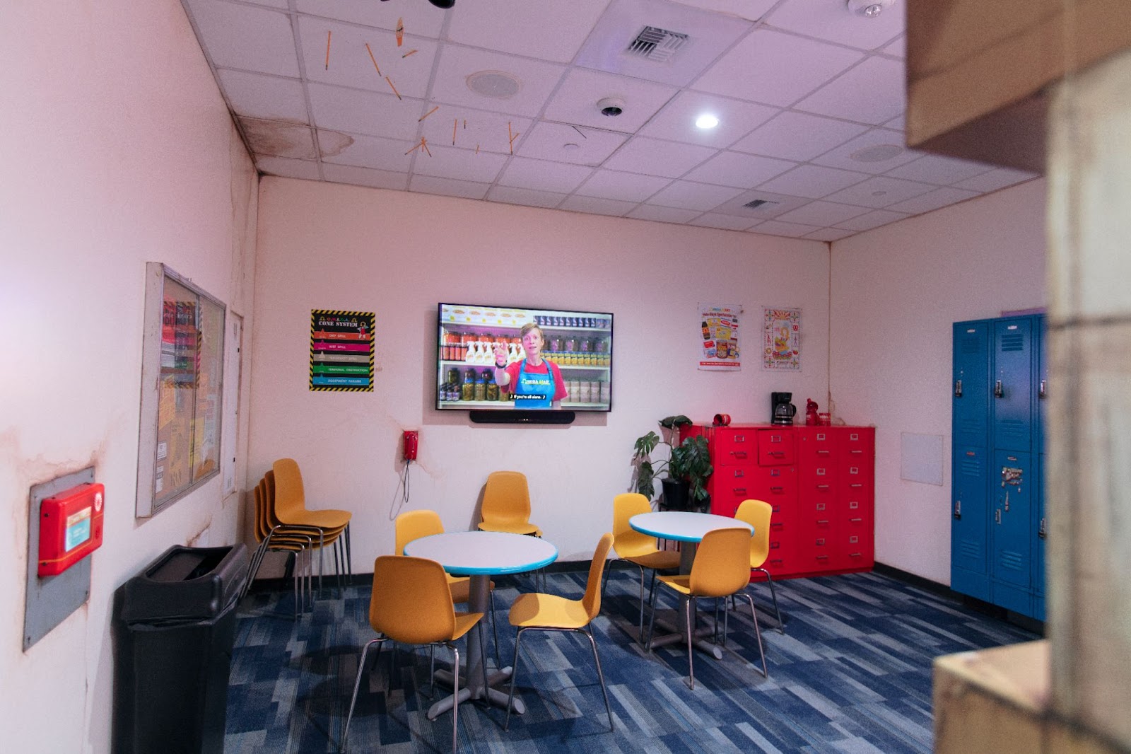 The employee breakroom has two tables in the center, each surrounded by three yellow chairs. There are more yellow chairs stacked in the corner and up against the wall. There’s a large tv on the wall, with an employee training video playing on loop. To the left of television is a cone system chart and to right of the tv is a bright red filing cabinet with an broken coffee pot on top. There are blue employee lockers along the wall near the filing cabinet. The ceiling has several water stains and there are pencils stuck on the ceiling as well.