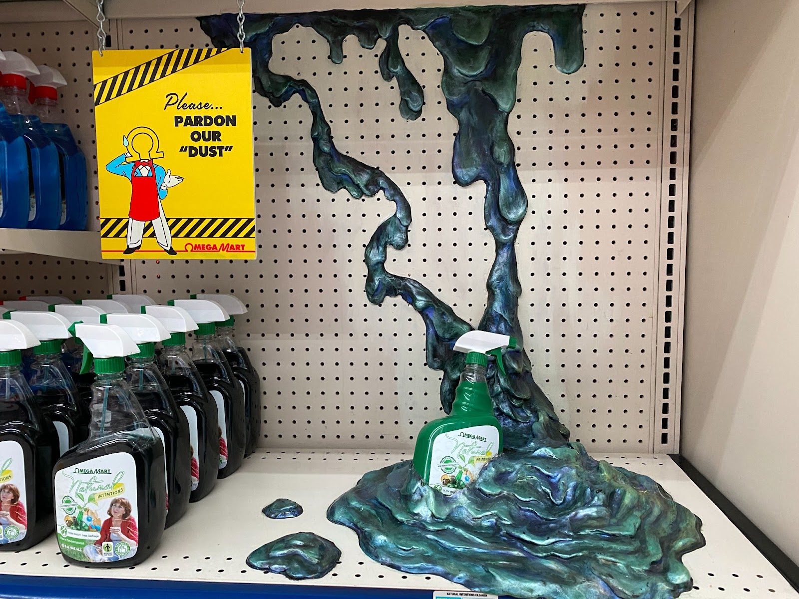 Blue and green goo oozes from a shelf above onto the bottom shelf. The goo has molded itself around a bottle of Natural Intentions household cleaner. There is a sign to the left featuring Mr. Omega looking apologetic and it reads “Please pardon our “dust”.