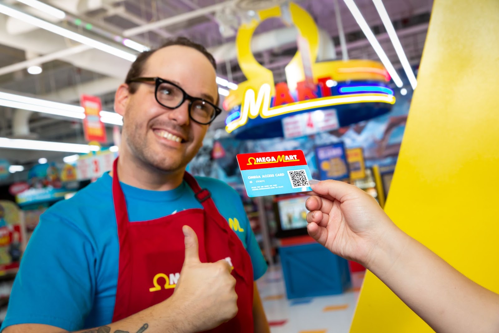 A man is the focal point of the image, he is wearing a blue Omega Mart shirt, with a red Omega Mart apron. He is wearing thick black glasses and smiling broadly while giving a thumbs up. There is an arm extended from the right side of the photo holding the Omega Access card.