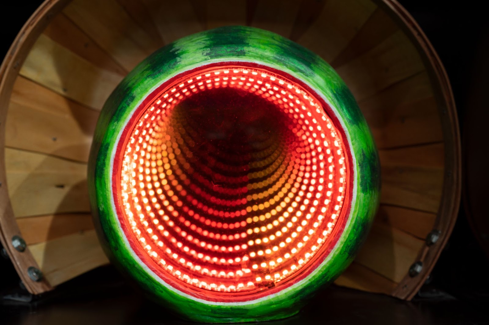 The left and the right side of the photo are dark and,m in the center, there is a large circular watermelon cut open to reveal a spiral of red and orange lights that seem to go on forever