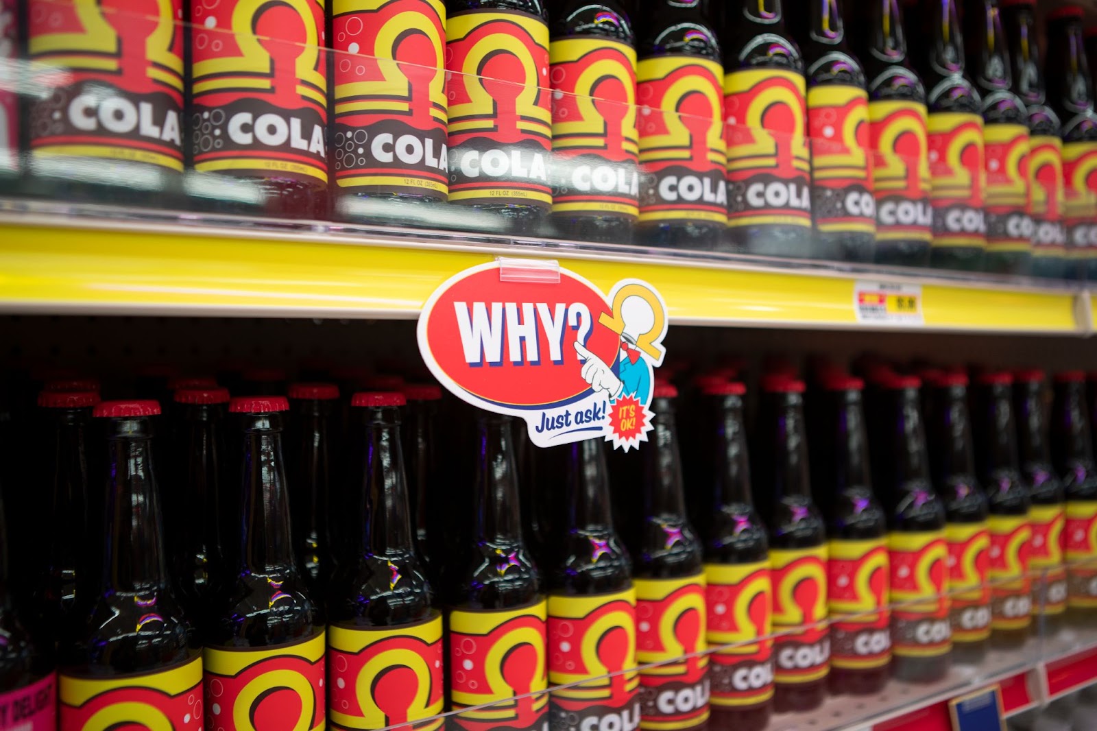 Omega Mart Cola lined up on the shelves. In the center of the image is an insert of the shelf of Mr. Omega pointing to a sign that reads “Why? Just ask! It’s okay!”