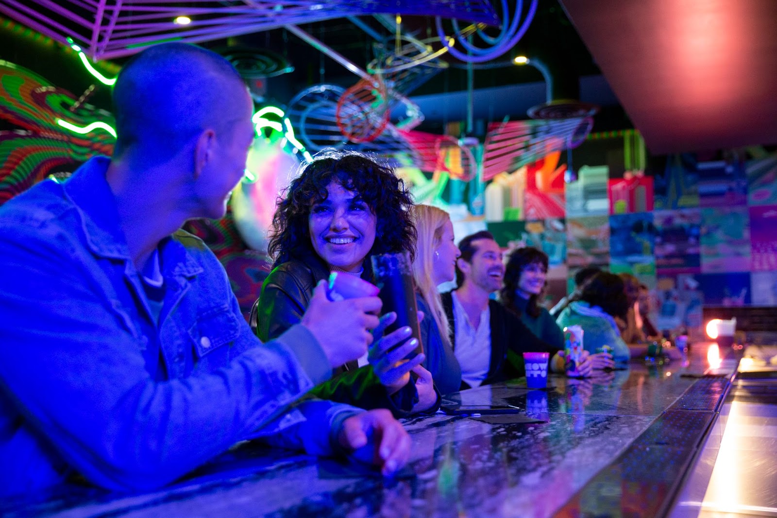 Guests sit at a bar counter with various beverages in front of them. In the forefront of the image, there is a man in a denim jacket holding his cup up in a cheers motion. The woman beside him is smiling and holding up her own drink to cheers with him.