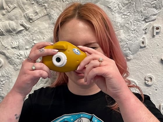 एक व्यक्ति सफेद पैटर्न वाली दीवार के सामने फोटोग्राफर की ओर आलू जैसा कैमरा रखता है।