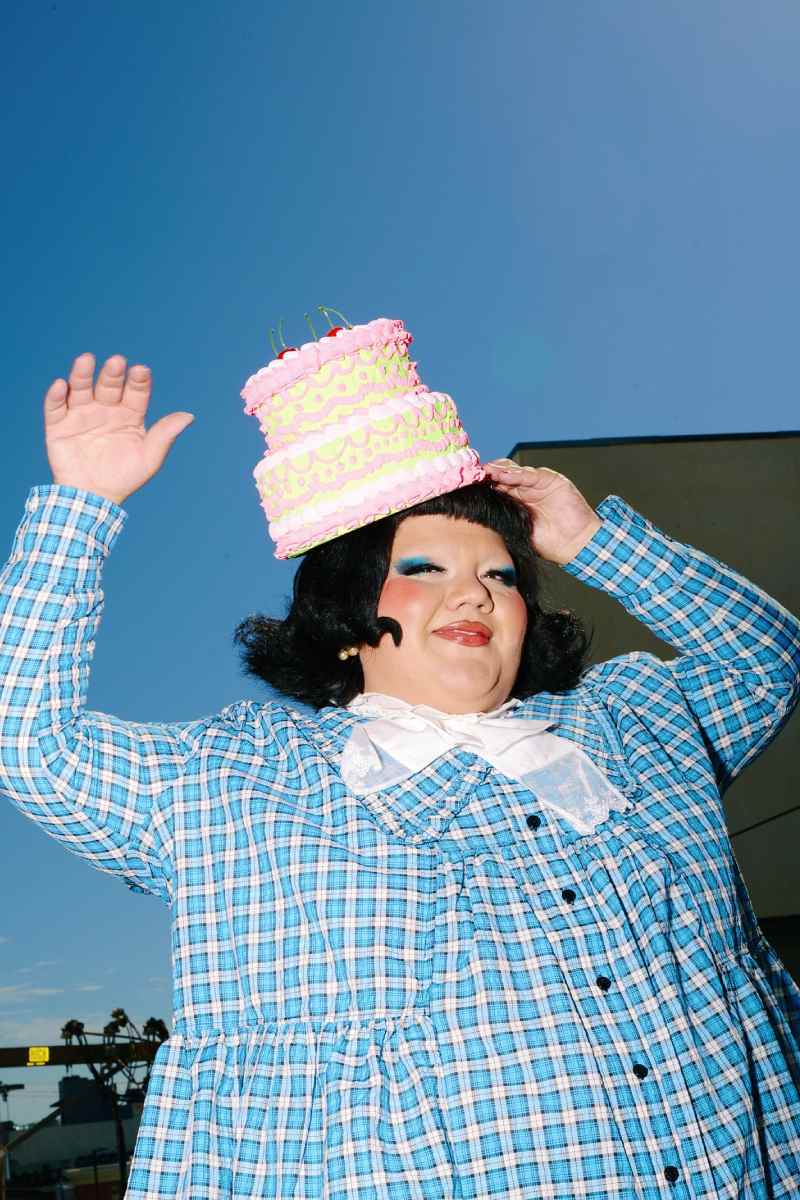 A person smiles at the camera with a short black bob, their hand holding a cake hat on their head.