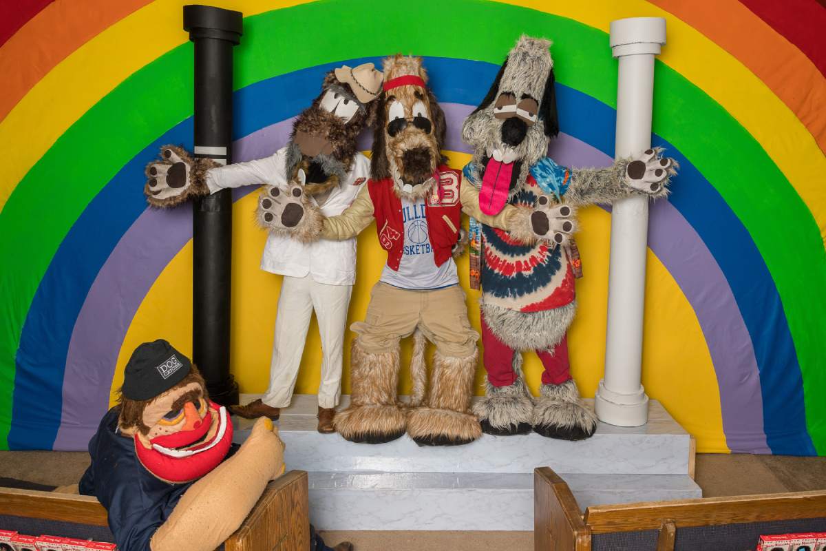 A group of dog like costumed figures stand on a small stage, another costumed humanoid figure sits in front of them. A painted rainbow is behind them.