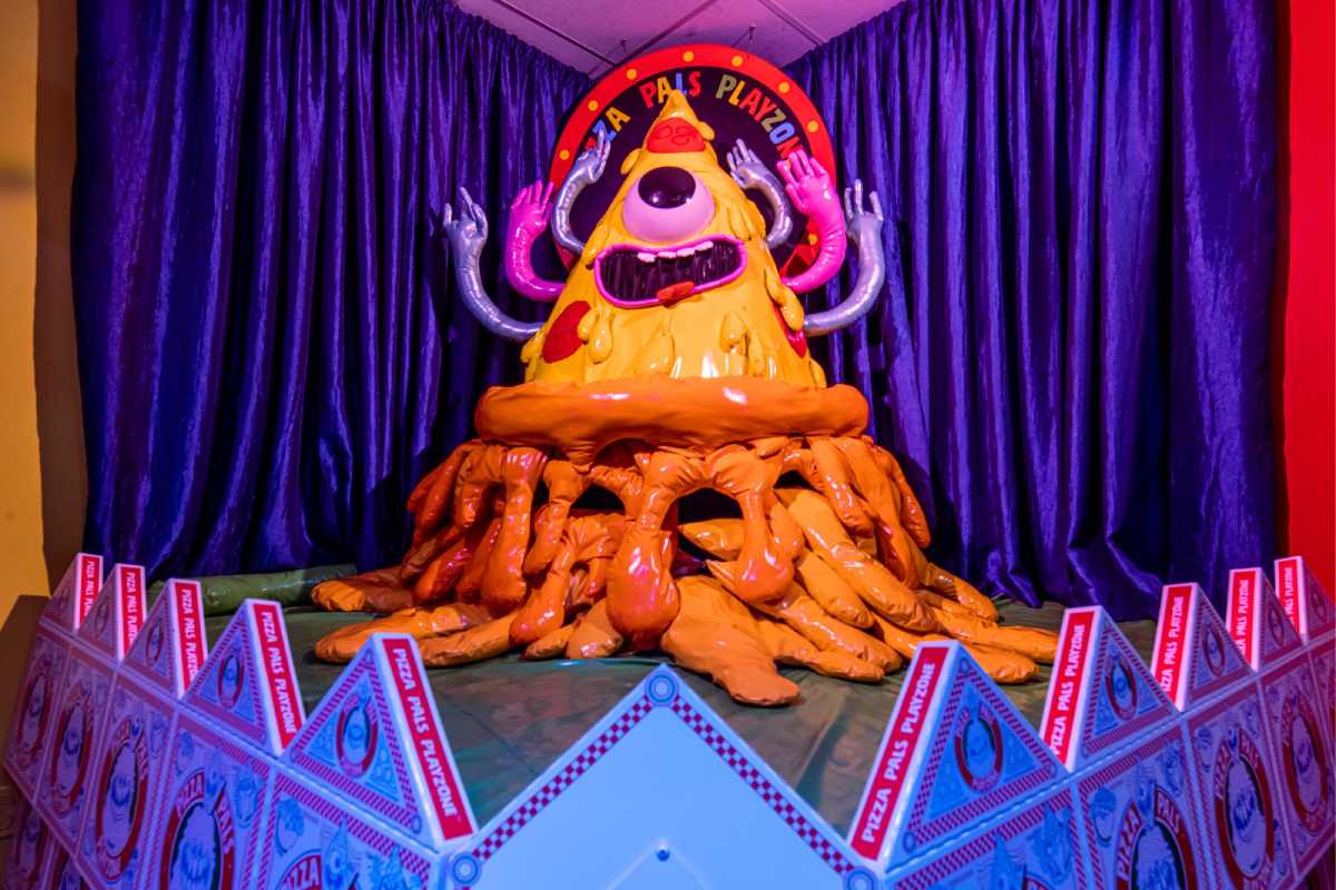 A pepperoni pizza costume stands in the center of a curtained stage with pizza boxes surrounding the edge.