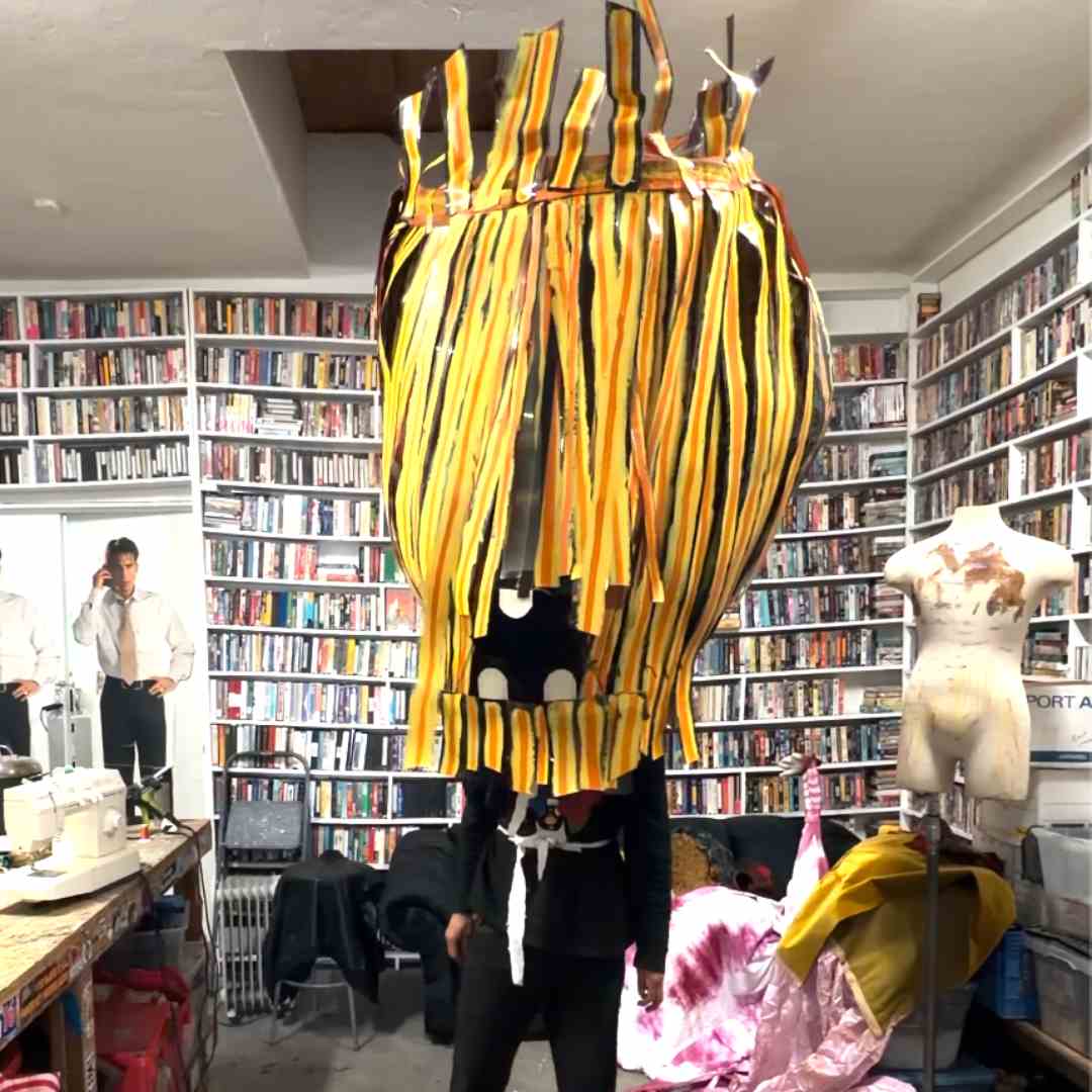 A person stands with a large headpiece on that’s made of yellow and orange film strips.