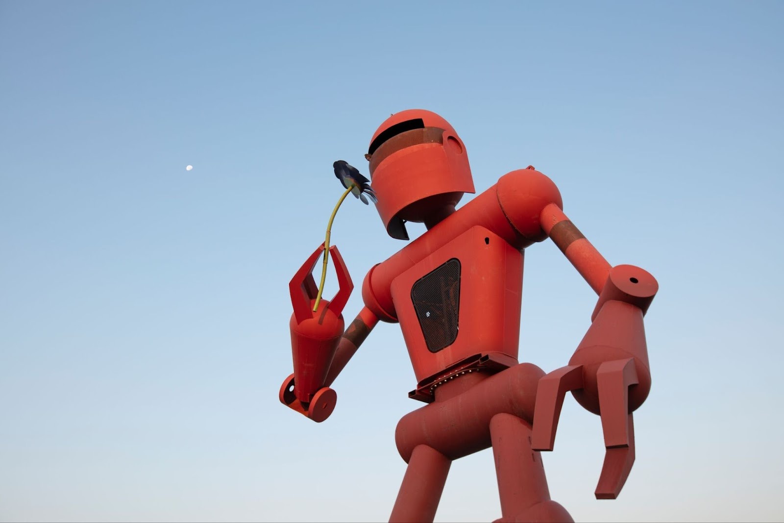 A large red robot smells a flower and a clear sky and moon is behind them.