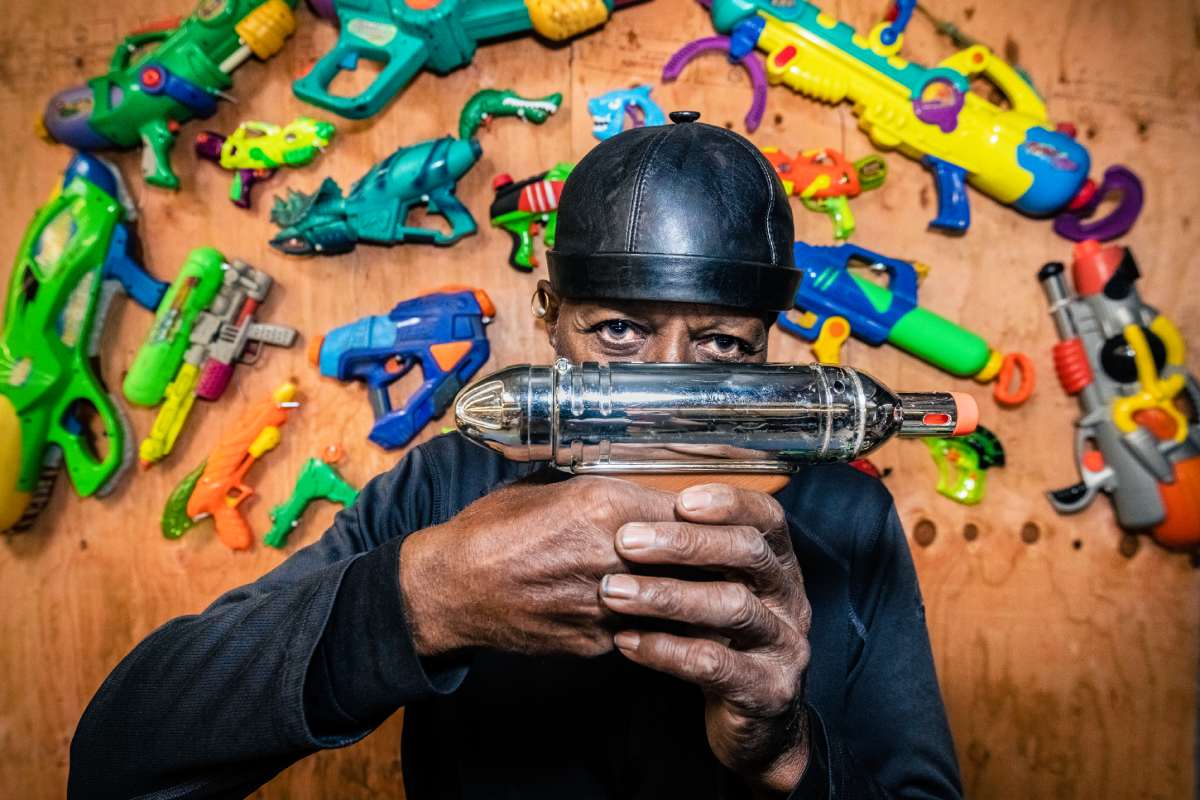A person in a black cap and long-sleeved shirt holds a metallic water toy with other colorful water guns behind them mounted on a plywood wall.