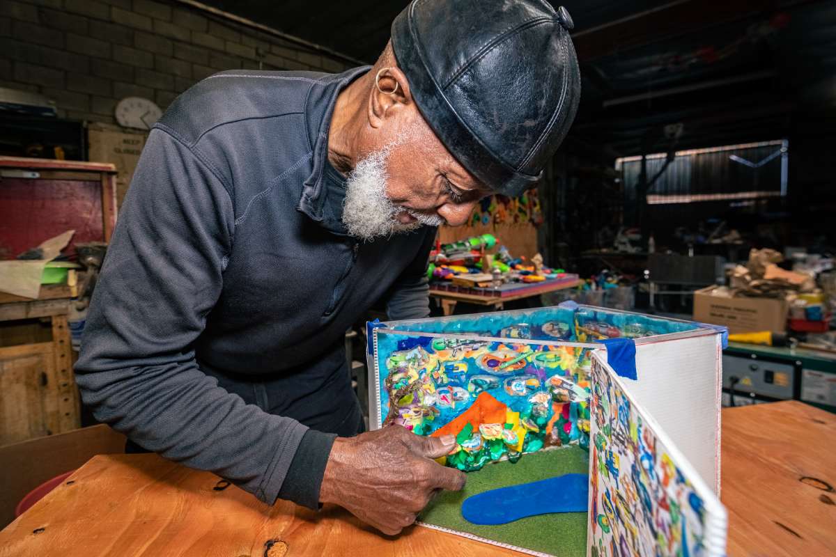 A person in all black clothing leans over a diorama with a colorful scene.