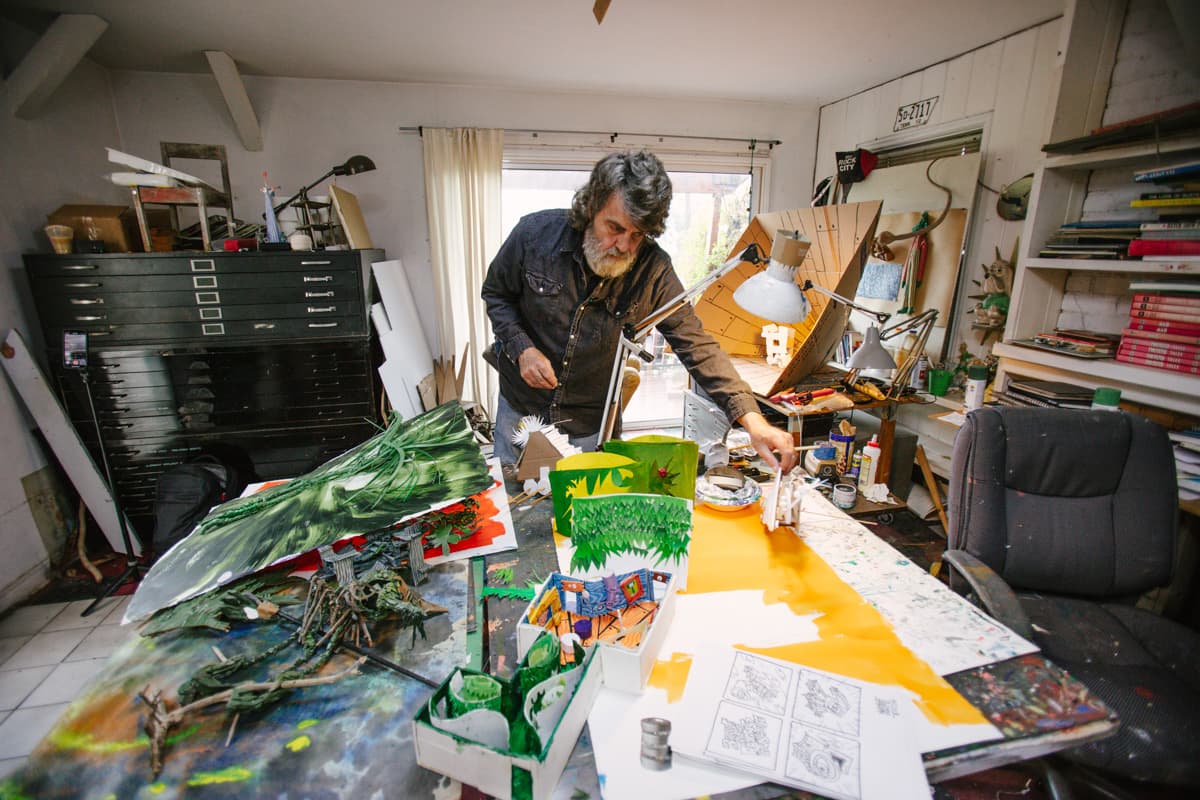 Artist Wayne White leaning over a table with several dioramas and models in his studio space
