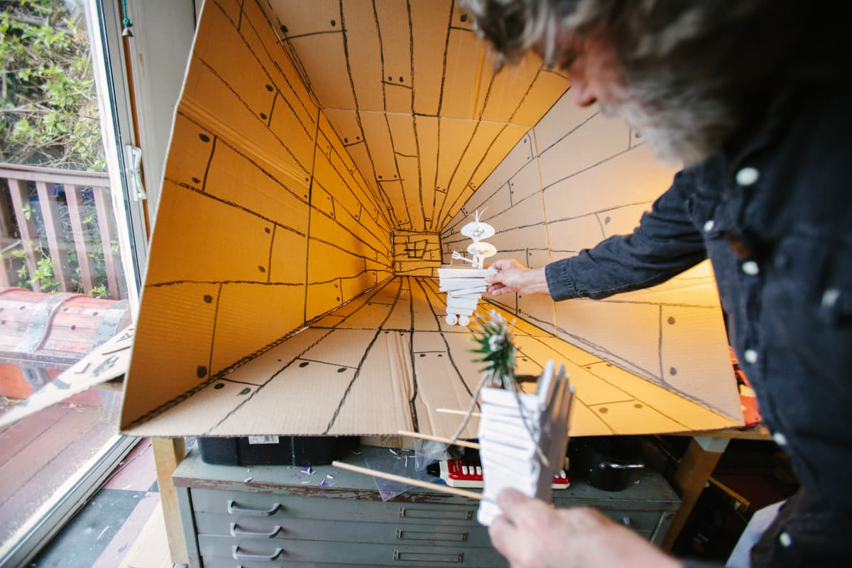 View of Wayne White holding a small model that looks like lightbulb shapes in a larger model with hand-drawn lines that look like wood planks