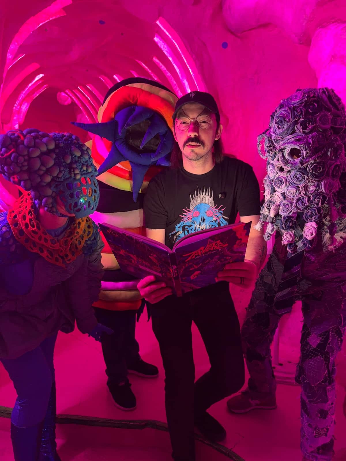 A human with an open book and other-dimensional beings surrounding the human. All are inside a pink-hued cave, maybe of a large ancient animal